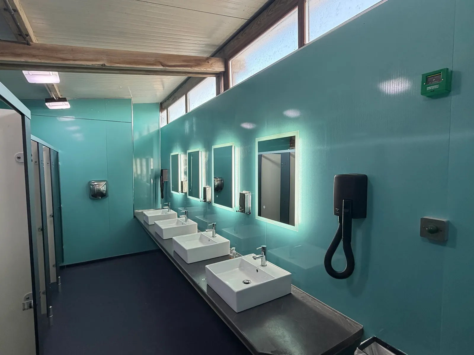 Inside the toilet and shower block: a long row of four white basins under lit mirrors on a turquoise wall, with a hairdryer on the wall and the cubicles on the left.