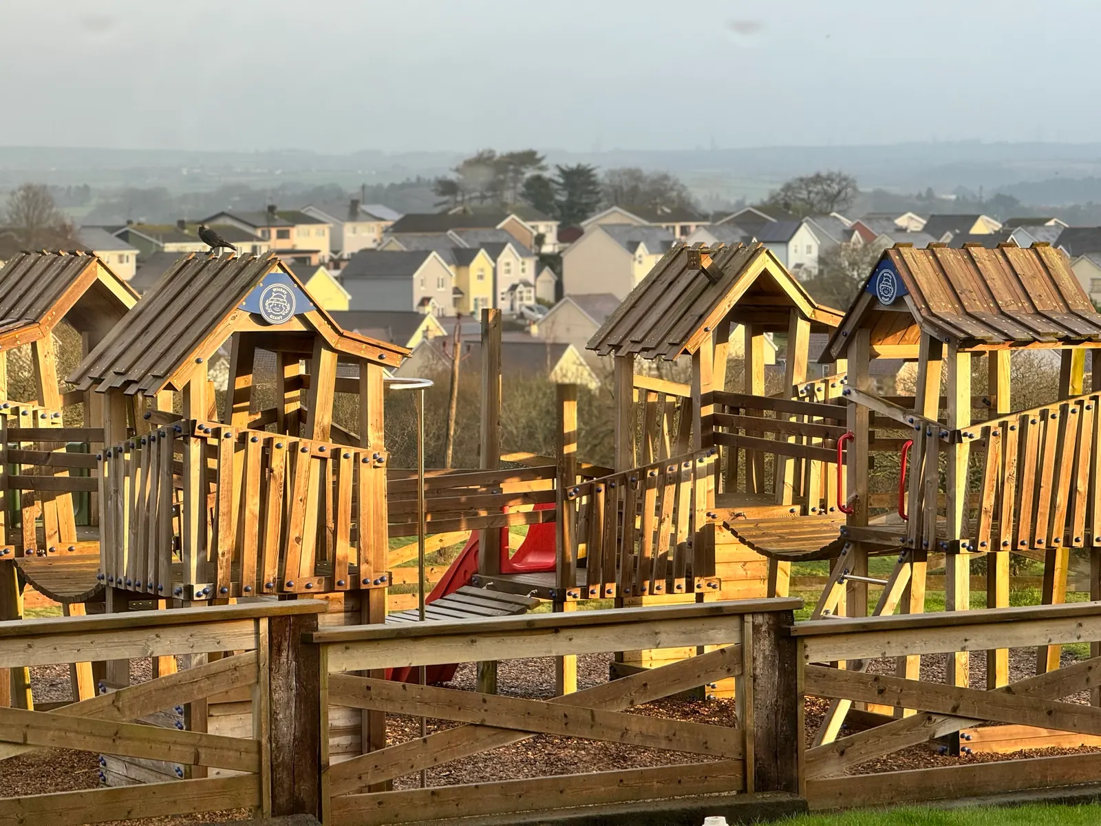 The wooden play towers in golden evening light, with their pointed roofs, a red slide and a wooden fence in front.