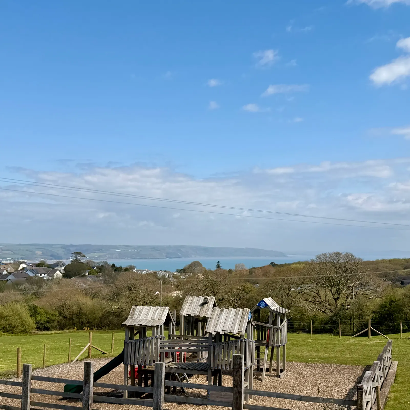 The wooden play area at Broadfield: play towers with slides on a bark floor inside a wooden fence, with green fields, trees and the sea of Carmarthen Bay behind under a blue sky.