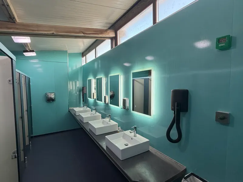 Inside the toilet and shower block: a long row of four white basins under lit mirrors on a turquoise wall, with a hairdryer on the wall and the cubicles on the left.